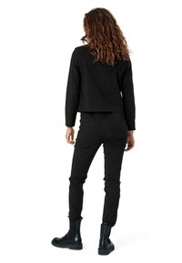 Kobieta z kręconymi włosami, ubrana w czarny żakiet, czarne spodnie i czarne botki za kostkę, stojąca tyłem na tle białym.