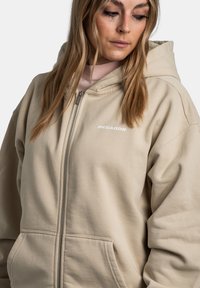 Beige zip-up hoodie van zacht materiaal, met een voorkanto pocket, koord in de capuchon en een wit "PEGADOR" logo op de linkerborst.
