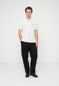 Πόλο μπλούζα σε εκρού βαμβάκι με γιακά και μαύρο λογότυπο. Συνδυάζεται με μαύρο cargo παντελόνι με πλαϊνές τσέπες και χαλαρή εφαρμογή.