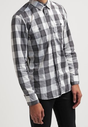 Camicia a maniche lunghe con motivo a scacchi in bianco e nero, colletto con bottoni, due tasche sul petto e polsini a contrasto. Il tessuto sembra leggero.