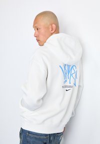 Sweatshirt branco com capuz e com uma gráfica e texto azuis nas costas. O tecido parece macio; apresenta um bolso canguru e punhos canelados.