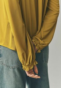 Långärmad blus i senapsgul, med samlade manschetter och ett luftigt tyg, ihopparad med högmidjade jeanstights.