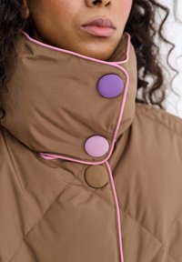Brązowa pikowana kurtka z wysokim kołnierzem, ozdobiona różowym przeszyciem i dwoma okrągłymi guzikami w kolorze fioletowym i brązowym. Gładka tekstura tkaniny.