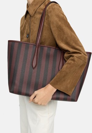 Osoba ubrana w brązową zamszową kurtkę i białe spodnie, niosąca dużą torbę typu tote z pionowymi ciemnoczerwonymi i czarnymi paskami oraz skórzanymi uchwytami.