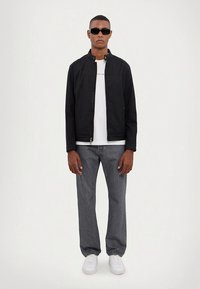 Must zip-up jakk valge särgi, hallide teksapükste ja valgete tossude kohal. Modell kannab musta päikeseprille, seisab sirgelt, käed küljel.