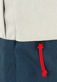 Bolso bicolor con una sección superior beige y un fondo azul oscuro. Incluye un cordón rojo con herrajes negros para el cierre. Tejido texturizado.