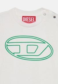 Balta medvilninė marškinėliai su dideliu žaliu Diesel logotipu ovalo formoje, su raudonu prekinio ženklo etikete ant kaklo ir dviem spaudėmis.