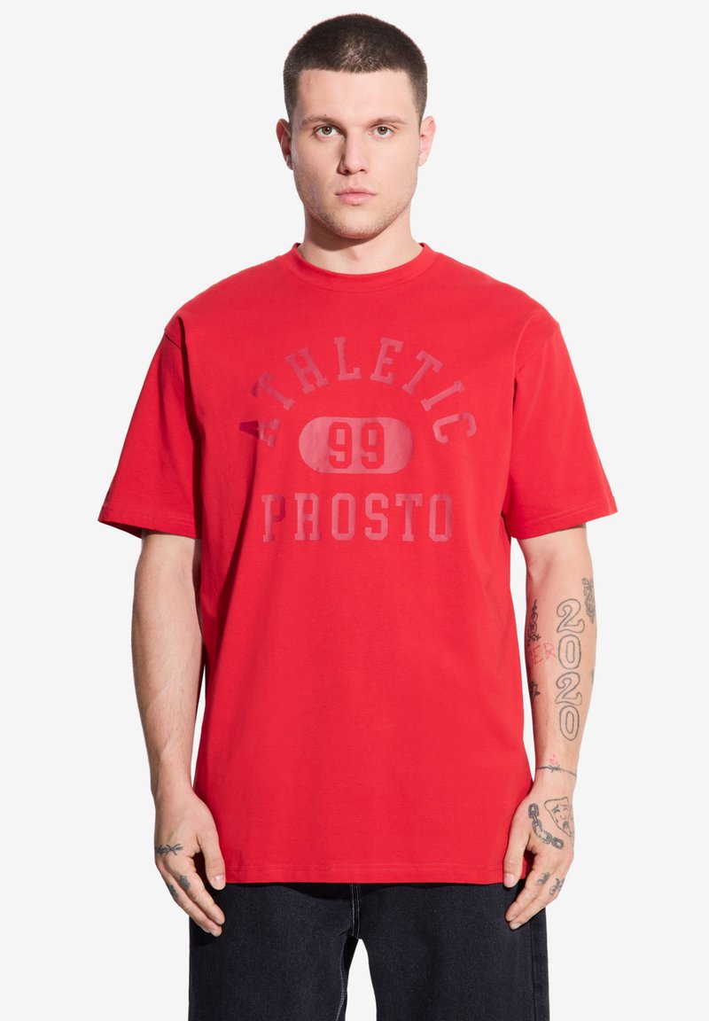 Czerwony bawełniany t-shirt z okrągłym dekoltem, krótkimi rękawami i nadrukowanymi grafikami z napisem "ATHLETIC 99 PROSTO" w kontrastowych kolorach.