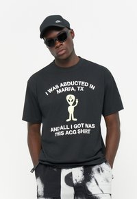 Svart bomulls t-shirt med vit text och en gul utomjording grafik. Kombinerad med svarta mönstrade shorts och en svart keps, med solglasögon.