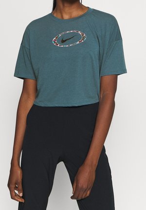 Person trägt ein taubenblaues, kurzes Nike-T-Shirt mit einem mehrfarbigen, floralen Oval um das schwarze Swoosh-Logo, kombiniert mit hoch taillierter schwarzer Hose.