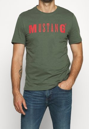 Mężczyzna w oliwkowo-zielonej koszulce z czerwonym napisem "MUSTANG", niebieskie dżinsy, prawa ręka w kieszeni, lewa ręka pokazuje tatuaż klucza wiolinowego.
