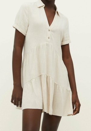 Robe courte en lin beige avec manches courtes, un col et deux boutons à l'avant, portée par une personne à la peau foncée.