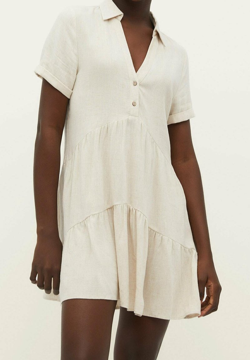 Robe courte en lin beige avec manches courtes, un col et deux boutons à l'avant, portée par une personne à la peau foncée.