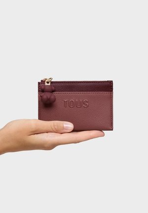 Hand, die einen burgunderfarbenen TOUS-Kartenhalter mit einem kleinen, bärenförmigen Reißverschlussanhänger vor einfarbigem Hintergrund hält.