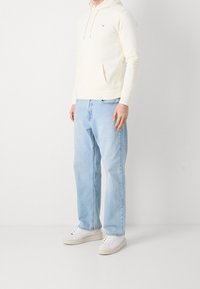 Sudadera crema clara con un bolsillo frontal y cordones; combinada con jeans rectos de color azul claro y zapatillas blancas.