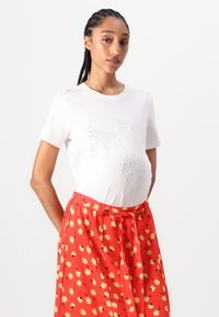 Witte t-shirt met korte mouwen, versierd met een tekstuur van een zilveren strik, gecombineerd met een vloeiende rode rok behangen met gele bloemenpatronen.