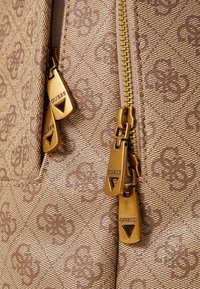 Sac en tissu beige avec un motif texturé, doté de fermetures éclair et de tirettes en métal doré affichant le nom de la marque "GUESS."
