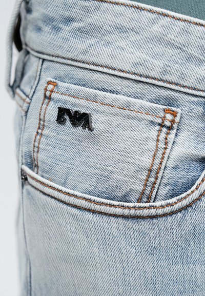 Lyseblå denimjeans baklomme med oransje søm og en liten svart metalllogo merket "EVA" festet til lommen.