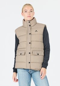 Pufferväst i beige med hög krage, svarta knappar, två framfickor och en svart logotyp på bröstet. Bärs över en långärmad tröja.