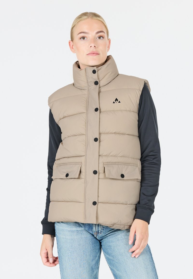 Pufferväst i beige med hög krage, svarta knappar, två framfickor och en svart logotyp på bröstet. Bärs över en långärmad tröja.