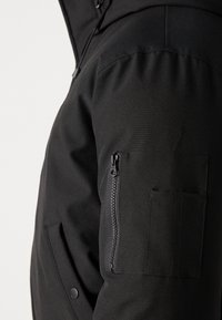 Black jacket featuring a zippered pocket on the sleeve, a textured fabric, and a hood, with visible stitching along the shoulder seam.