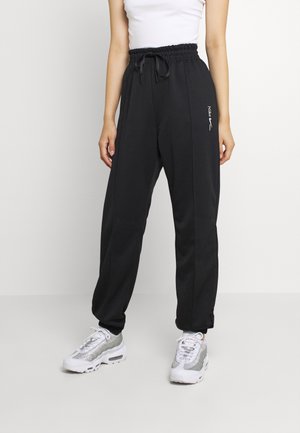 Osoba nosząca czarne dresy Nike z elastycznym pasem i ściągaczami, w połączeniu z białymi i srebrnymi butami sportowymi oraz białą koszulką bez rękawów.