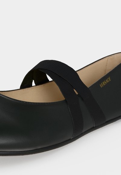 Chaussure plate en cuir noir avec des brides élastiques croisées et une doublure intérieure beige, avec un marquage doré Versace à l'intérieur.