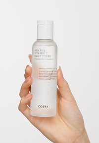 Bouteille transparente givrée avec un capuchon blanc ; étiquetée "Toner Quotidien AHA BHA Vitamine C" avec du texte et des ingrédients en noir et accents aqua.