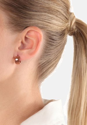 Nahaufnahme des Ohrs einer blonden Frau mit einem kleinen, polierten Ringohrring aus Roségold, die Haare zu einem Pferdeschwanz gebunden, vor einem schlichten Hintergrund.