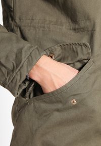 Olivgrön jacka med en framficka som har en guldfärgad tryckknapp. Tyget är slätt med en något strukturerad yta.