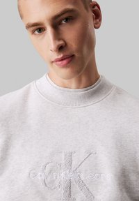 Sudadera de cuello redondo en gris claro, hecha de tela suave, con un logotipo bordado "CK" y el texto "Calvin Klein Jeans" en el frontal.