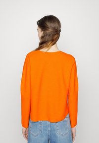 Heller orange gestrickter Pullover mit langen Ärmeln und rundem Ausschnitt; endet auf Taillenhöhe; getragen mit hellblauen Denim-Jeans.