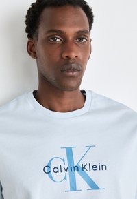 Camiseta de algodón azul claro con el logo "Calvin Klein" en letras azul oscuro y azul claro, cuello redondo, mangas cortas y textura suave.