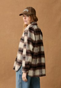 Kostýmová košeľa z vlnenej zmesi v krémovej a tmavej burgundskej farbe, s golierom, dlhými rukávmi a gombíkovými manžetami, doplnená o svetlomodré džínsy.