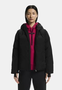 Fekete pufferszövet kabát kapucnival és cipzáros elülső résszel, rózsaszín pulóver fölött viselve. Szellős fazonja és bordázott mandzsettái vannak.