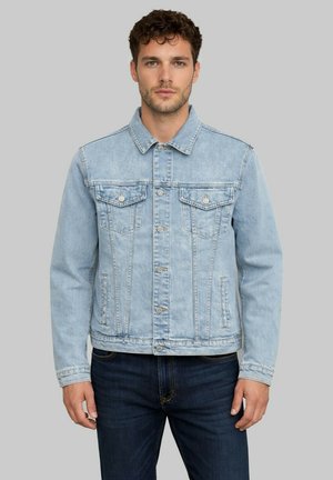 Homme portant une veste en jean bleu clair avec fermeture à boutons et un jean bleu foncé, debout devant un fond gris uni.