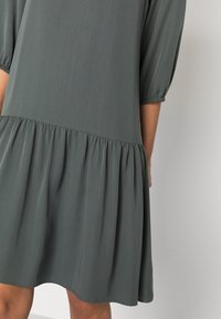 Vestido de manga corta en tela verde suave con falda acampanada, que presenta un dobladillo redondeado y sutiles fruncidos en las mangas y la cintura.