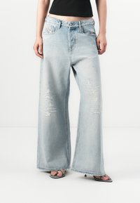 Personne portant un jean large déchiré bleu clair, un haut noir court et des sandales à talons hauts à brides sur fond blanc.