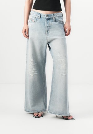 Person wearing light blue distressed wide-leg jeans, black cropped top, and strappy high-heeled sandals against a white background.