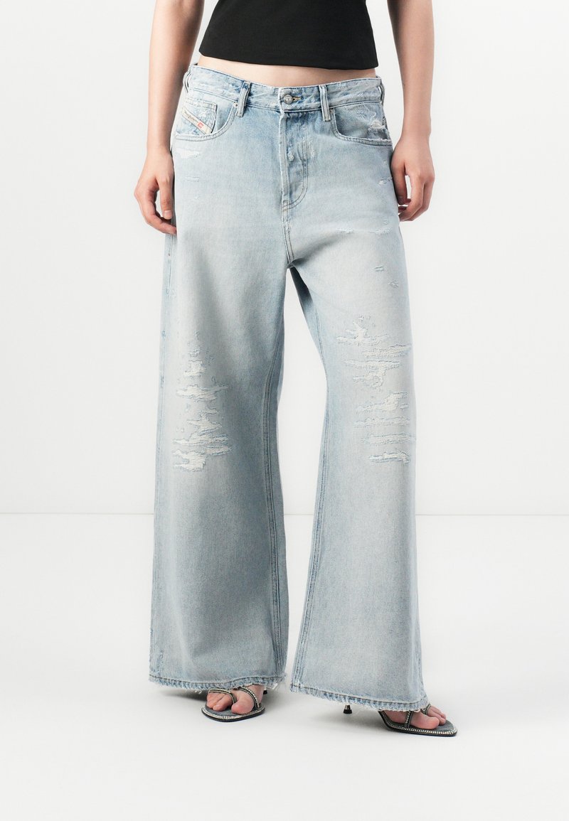 Personne portant un jean large déchiré bleu clair, un haut noir court et des sandales à talons hauts à brides sur fond blanc.