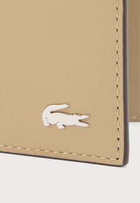 Bež kožni kutak novčanika s vidljivim šavovima i metalnim srebrnim detaljem u obliku krokodila. Glatka tekstura s minimalističkim dizajnom.