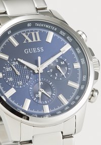 Montre en acier inoxydable argenté avec un cadran texturé bleu, des accents argentés, des fonctions chronographe et un tachymètre autour de la lunette.