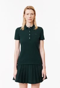 Polo shirt verde scuro con maniche corte, caratterizzato da un colletto e una chiusura a bottoni. Abbinato a una gonna a pieghe verde scuro.