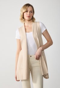 Beige ärmlös väst med öppen front, mjuk struktur, två framfickor, bärs över en vit t-shirt och ljusa byxor.