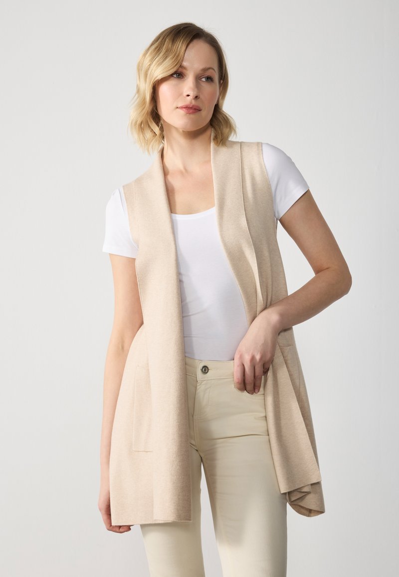 Beige ärmlös väst med öppen front, mjuk struktur, två framfickor, bärs över en vit t-shirt och ljusa byxor.