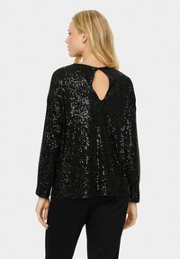 Top lungo a maniche con paillettes nere, con scollatura rotonda e apertura a chiave sul retro. La superficie testurizzata e lucida aumenta l'interesse visivo.