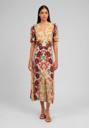 Femme aux longs cheveux foncés portant une robe midi à manches courtes et col en V, à motifs floraux rouges, jaunes, verts et marron, et des sandales à brides beige.