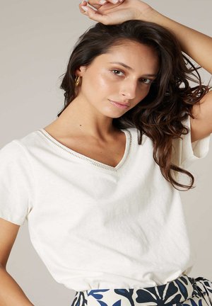 Jeune femme aux cheveux ondulés et foncés, portant des boucles d'oreilles dorées en forme d'anneau, un t-shirt blanc à col en V et une jupe à motifs, posant avec un bras levé.