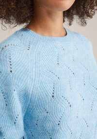 Lichtblauwe gebreide sweater met een golfpatroon, geribbelde halslijn en open steekdetails, die een textuurachtig uiterlijk geven.