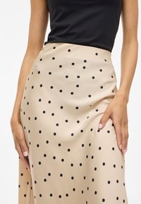 Bež krilo s črnimi pikami, gladka tekstura, do kolen, prilegajoč pas, roka naslonjena na tkanino za podporo.